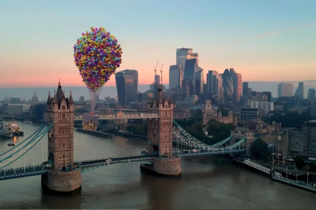 An aerial view of London's Tower Bridge at sunset, with the cluster of colourful balloons from the Pixar film Up, floating above the bridge and cityscape.