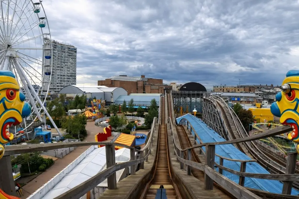 View from the top of Dreamland’s Scenic Railway, looking down wooden tracks over an amusement park with a Ferris wheel, colorful rides, and a cloudy sky.
