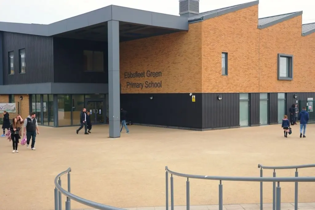 Brick and black-clad exterior of Ebbsfleet Green Primary School, with parents and children walking across a large open courtyard in front of the main entrance.