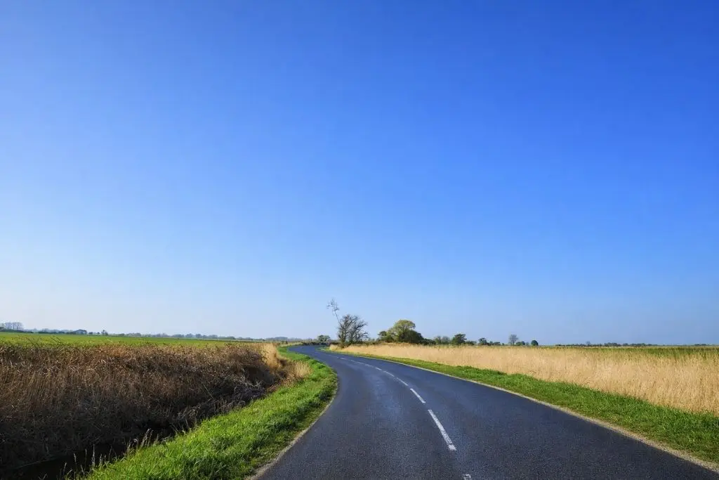 A paved rural Romney Marsh road curves to the right, with grass and fields on both sides and a clear blue sky above